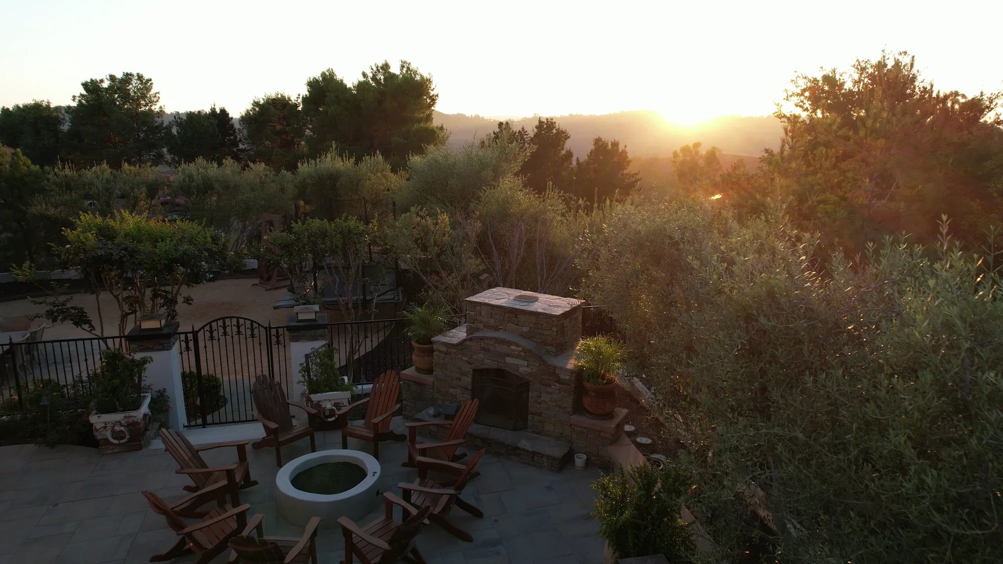 Elevated view of a fire bowl, wooden chairs and stone fireplace among planting at sunset.