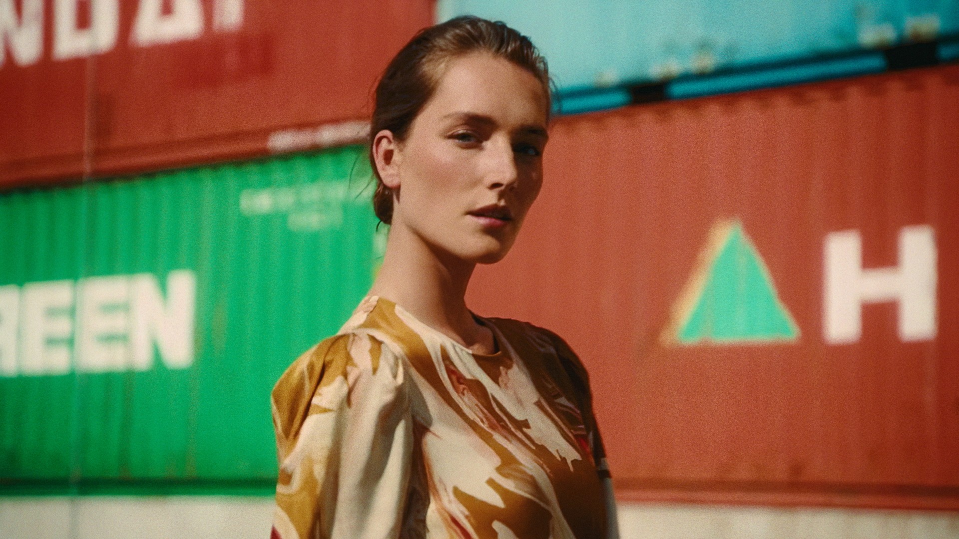 A woman in a gold printed dress in front of red and green shipping containers, Lafayette 148 New York