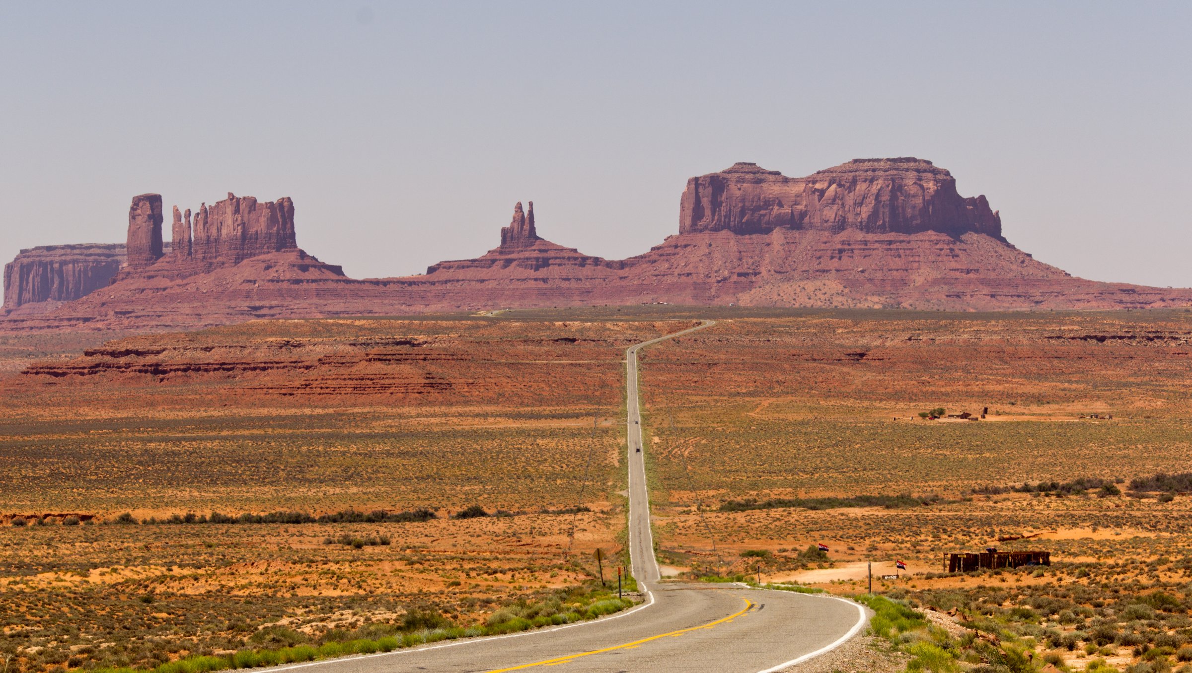 A long straight two-lane highway running toward the buttes of Monument Valley, the road narrowing to a point between red desert and a pale sky.