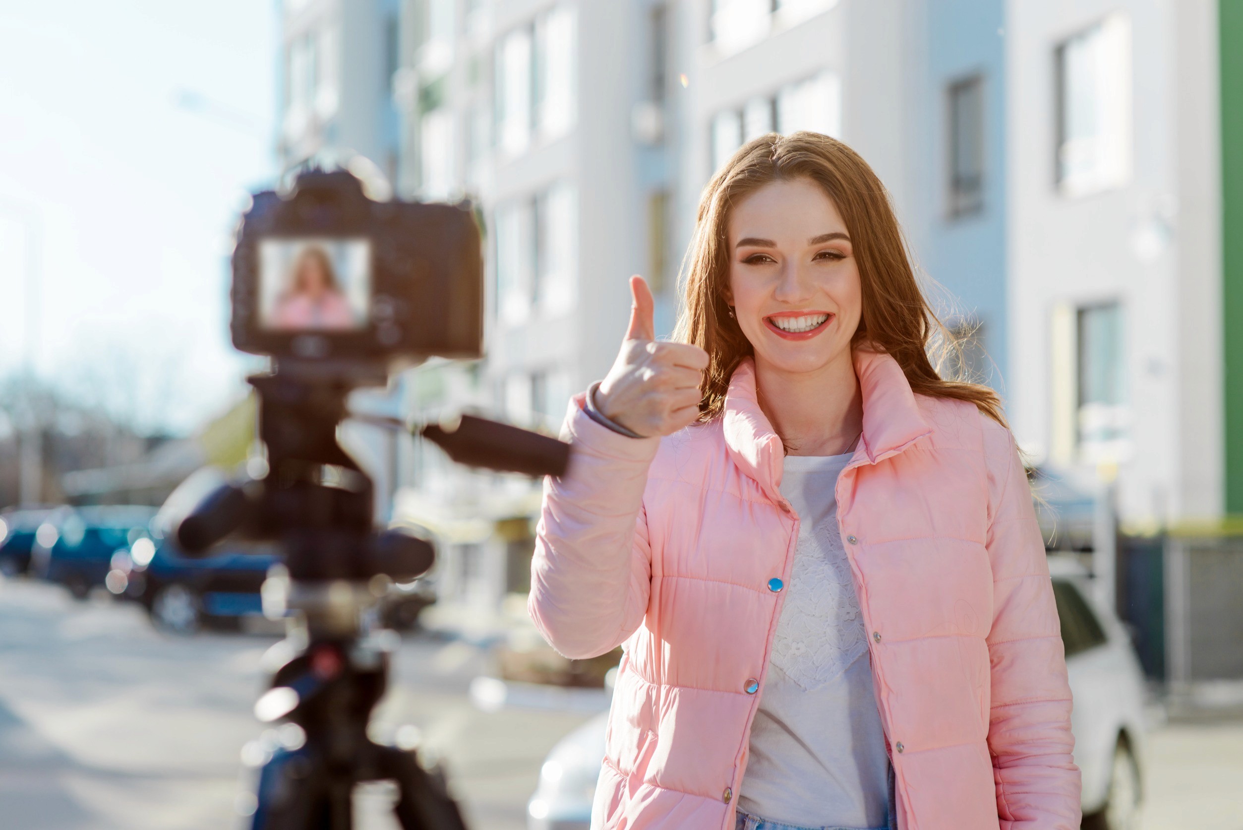 A young woman influencer filming content outdoors, smiling and giving a thumbs-up gesture to the camera. She is wearing a pink puffer jacket and standing in a sunny urban environment, with buildings and cars in the background. The camera is mounted on a tripod, capturing her positive and engaging expression.
