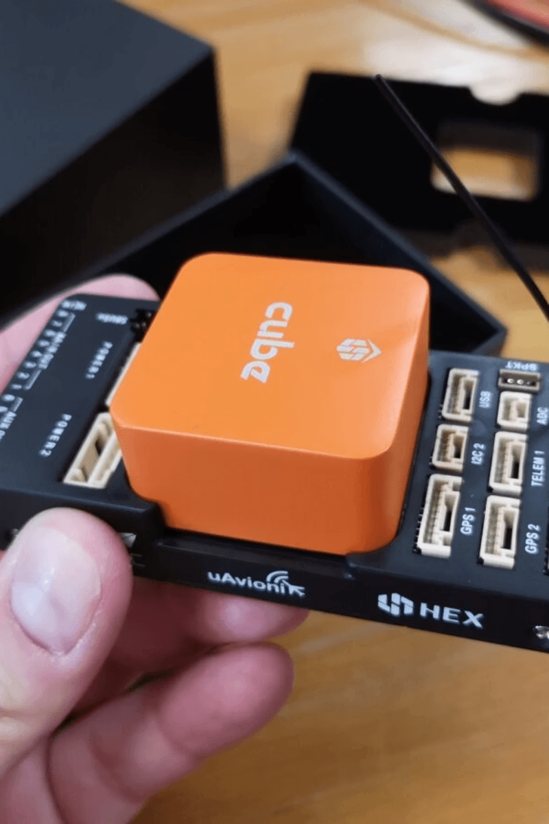 An orange flight controller module labeled ‘Cube’ is held in someone’s hand above a black autopilot circuit board with multiple connectors.