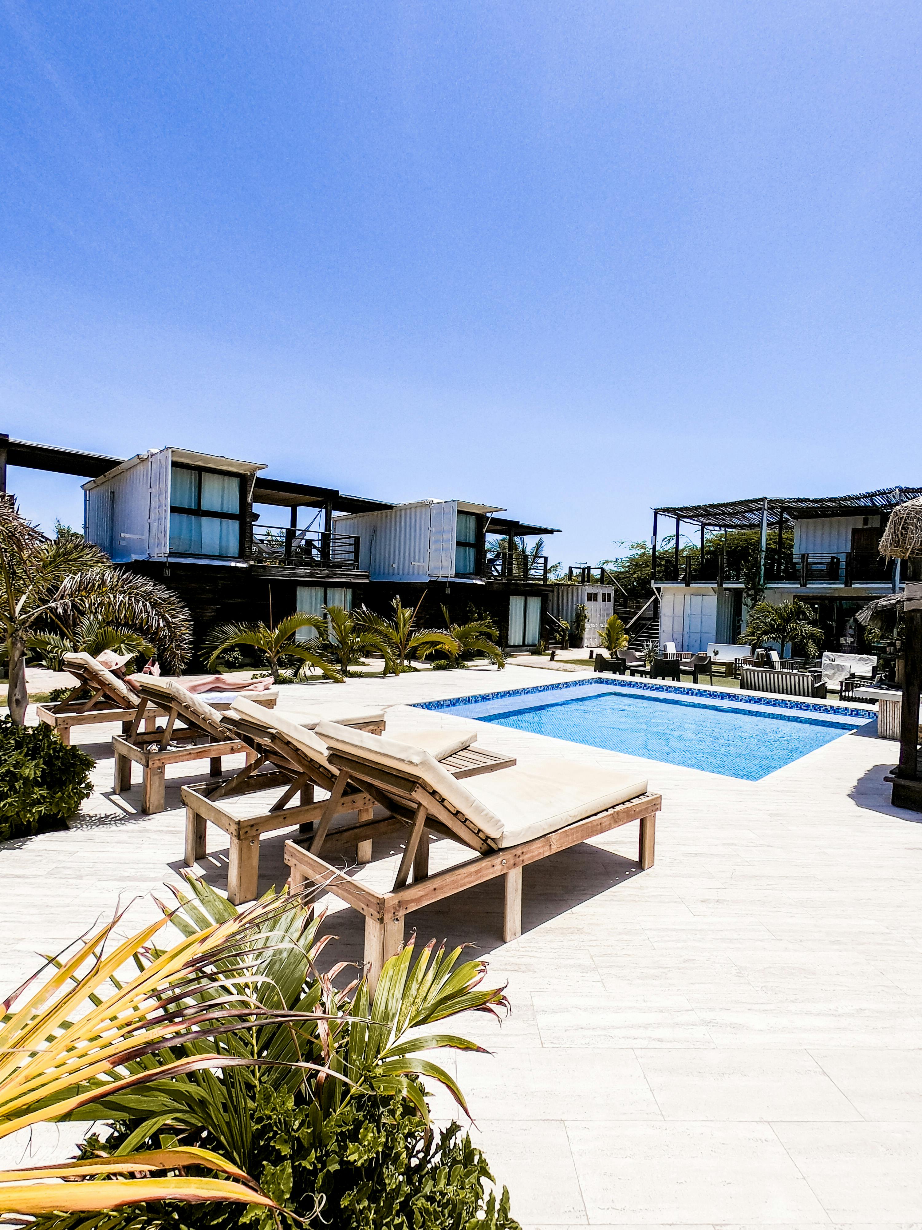 Stylishly arranged wooden sun loungers surround a sparkling blue swimming pool in a modern outdoor resort setting, featuring contemporary architecture and lush greenery under a clear blue sky.
