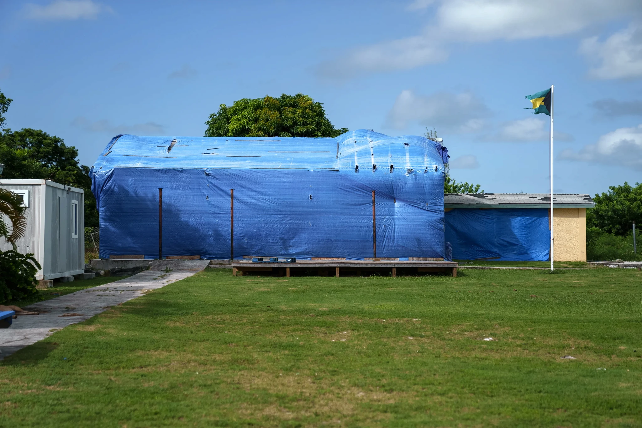 A building covered in a large blue tarp next to a small white structure. A flagpole with a green, yellow, and black flag stands on the right. The scene is set on a grassy field under a partly cloudy sky.