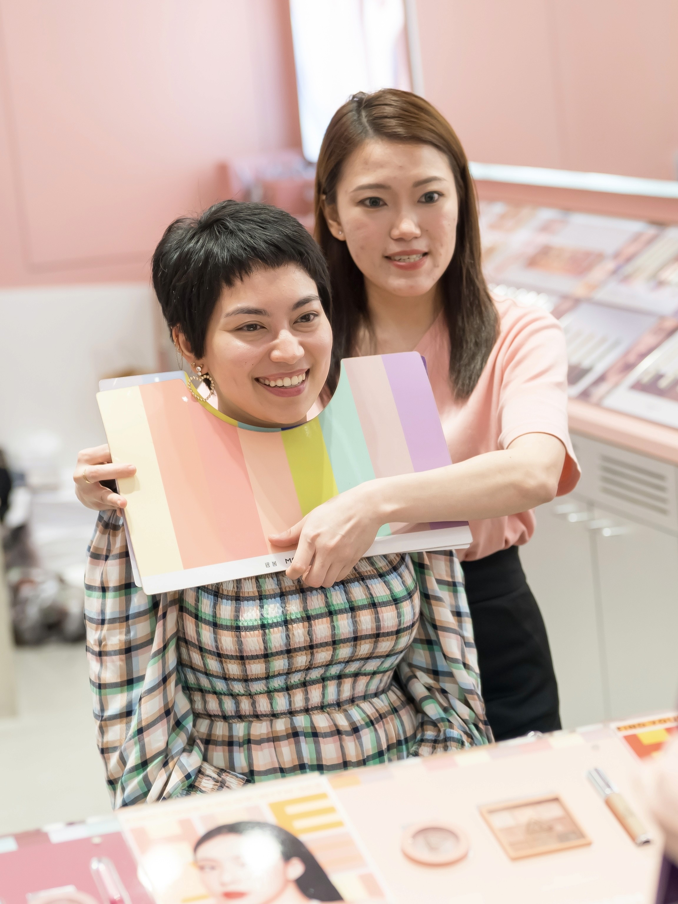 A stylist assists a client by holding a color palette next to her face, with various beauty products displayed on a table in a vibrant and modern setting.