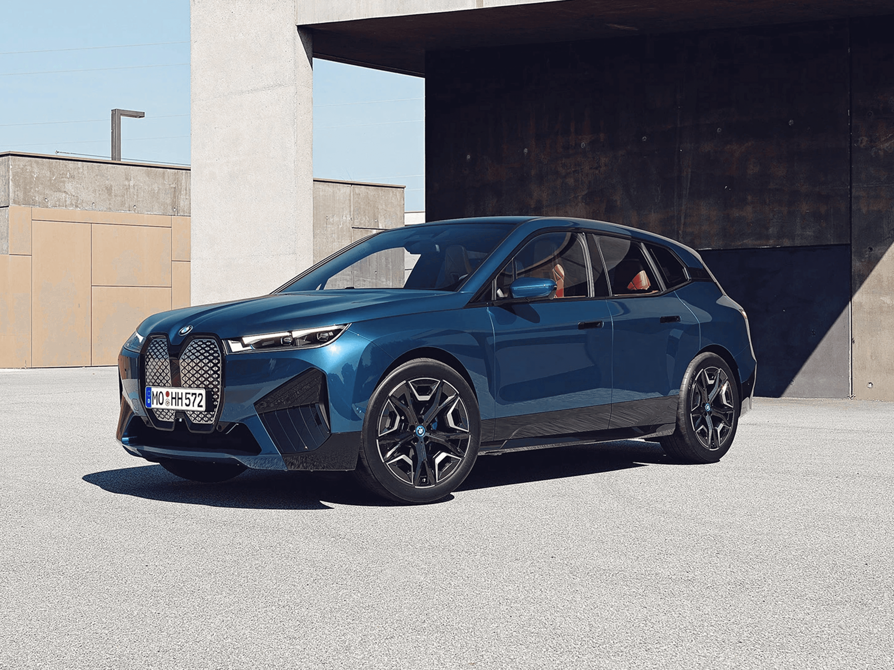 Rendering di suv elettrico BMW iX visto di tre quarti frontalmente, con carrozzeria blu ed interni in pelle in ambiente architettonico moderno.