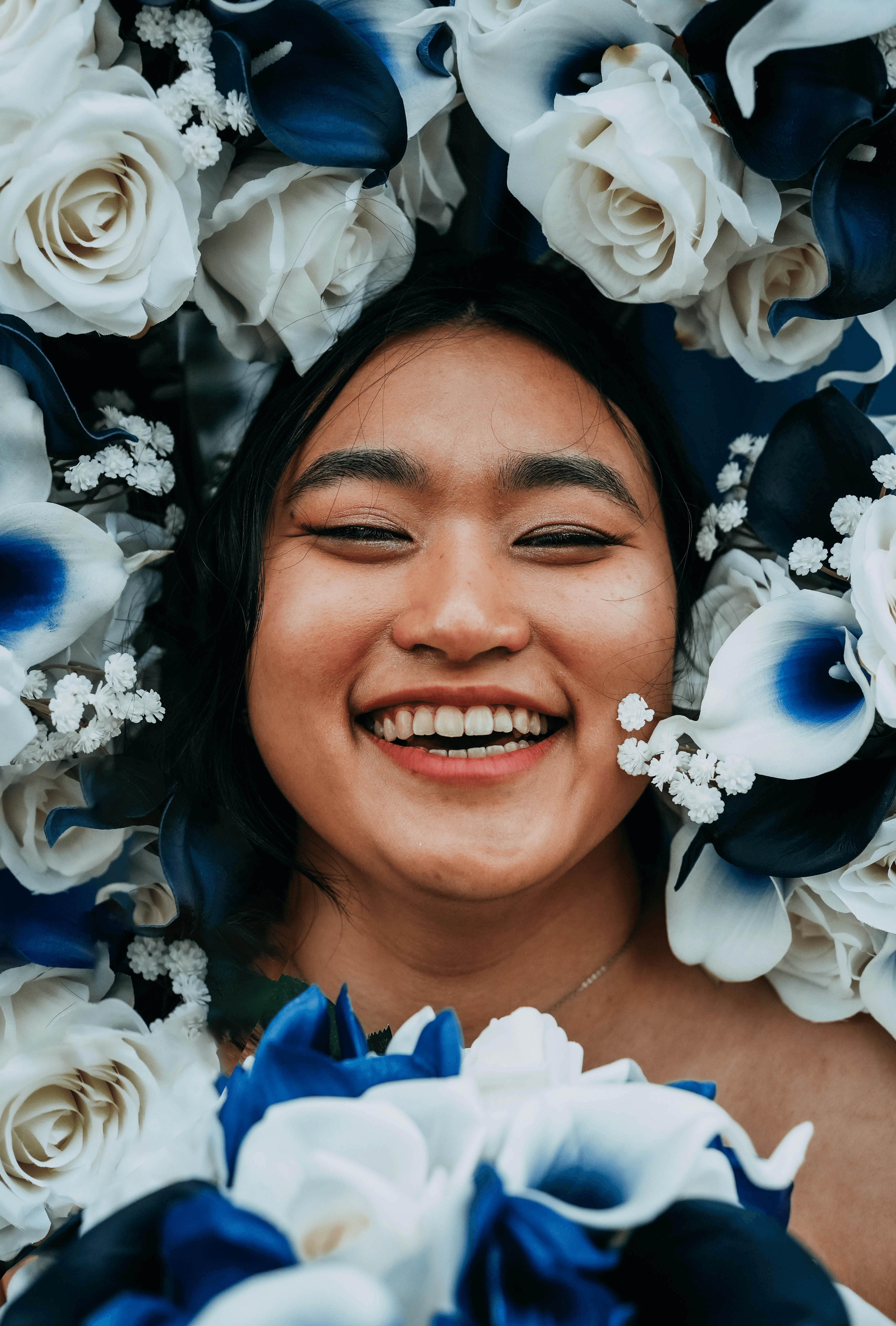 A joyful, close-up portrait of the bride laughing, with her eyes crinkled in happiness. Her face is surrounded by a frame of her white and blue wedding flowers.