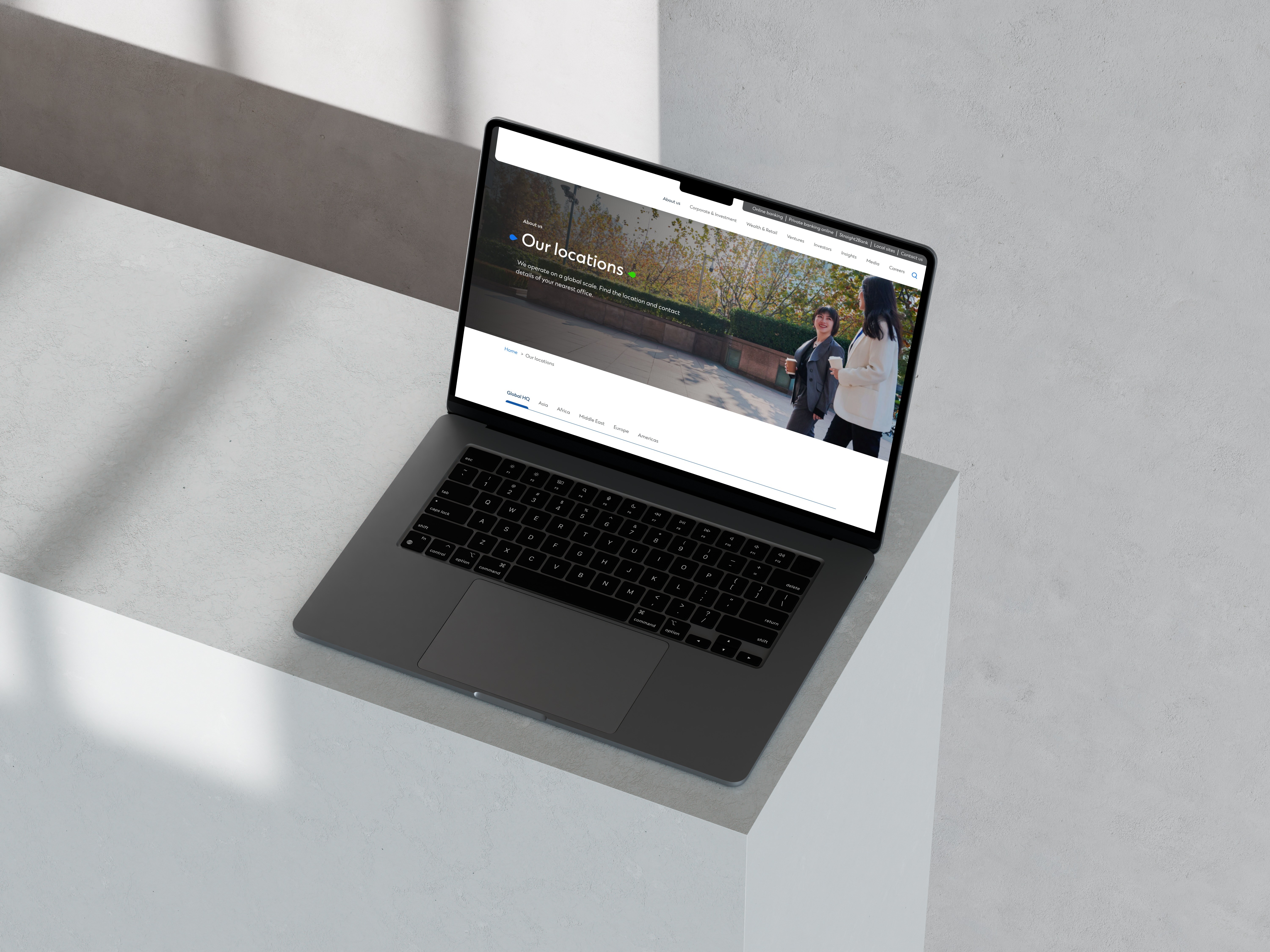 Un MacBook Pro est posé sur une surface blanche. L'écran affiche la page de localisation d'une entreprise avec une mise en page épurée. L'en-tête de la page "Nos emplacements" inclut du texte sur la recherche de bureaux à proximité. Le site a un menu simple avec des sections pour les services bancaires, les investissements et les informations sur l'entreprise. La lumière naturelle projette une ombre sur l'installation minimaliste.