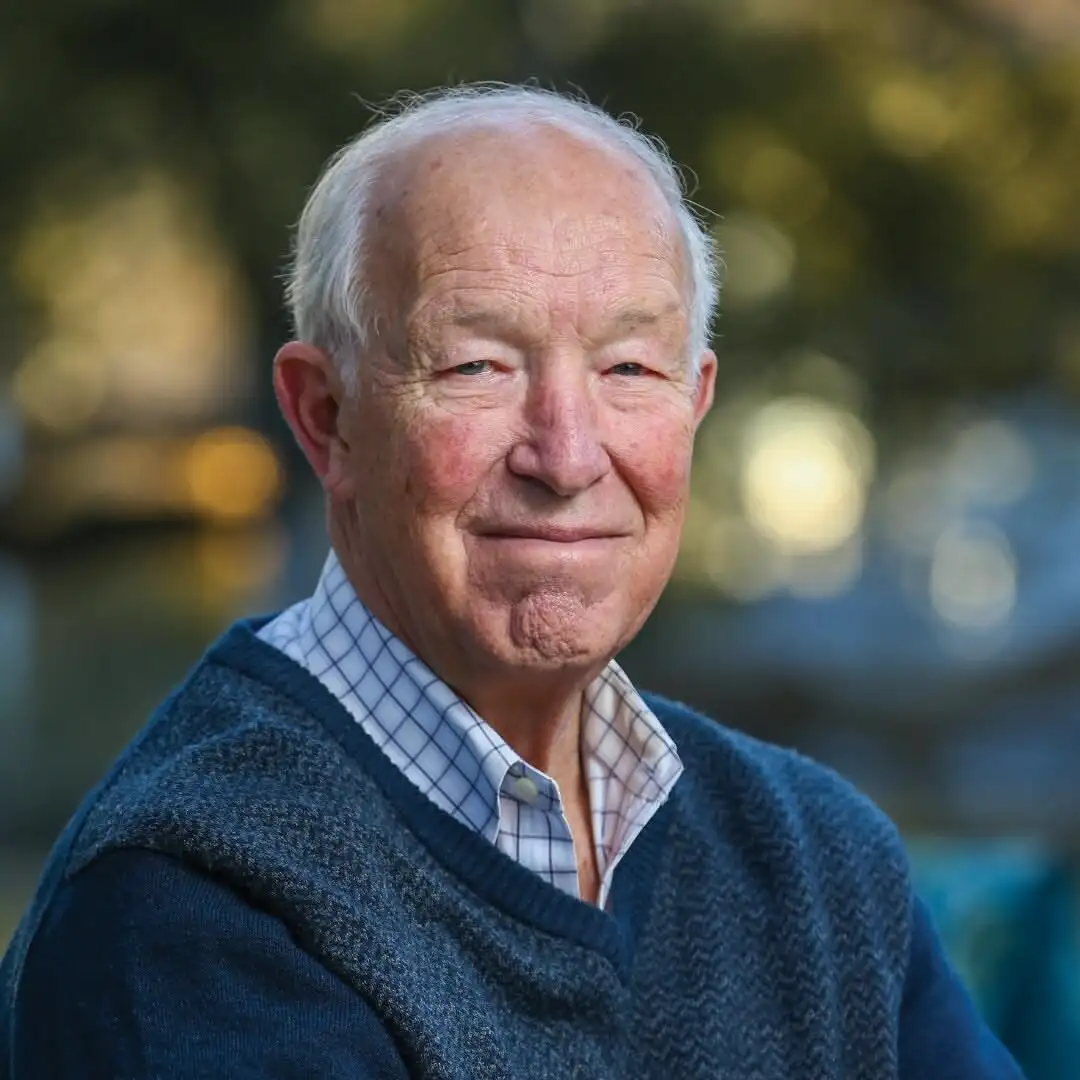 Elderly man with white hair wearing a navy sweater over a plaid shirt, smiling outdoors with blurred greenery behind him.