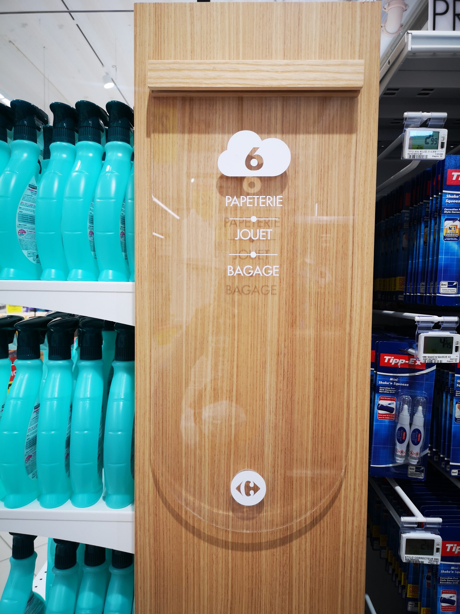 Panneau de signalisation dans un magasin, sur un panneau en bois, indiquant la section 6 avec les catégories suivantes : papeterie, jouets et bagages. À proximité se trouvent des bouteilles bleues et des produits de correction.