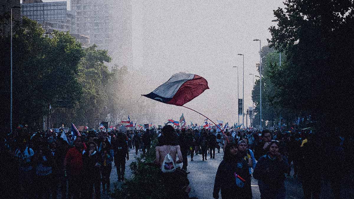Fotografía de decenas de personas que están marchando por la Alameda mientras una de ellas sostiene una bandera chilena, en la columna de opinión "Maldito infierno" de IdeaPaís. 