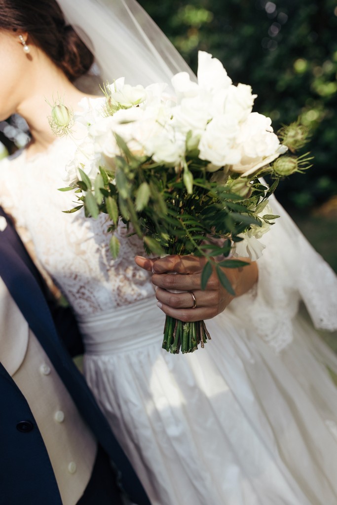 Sposa con bouquet di rose bianche e verdure, abito in pizzo bianco, luce naturale su villa privata