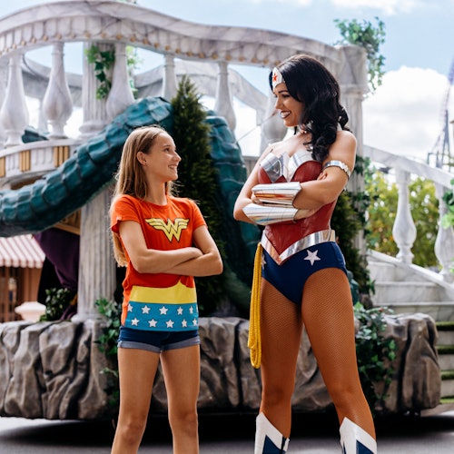 Een meisje in een Wonder Woman T-shirt glimlacht naar een vrouw verkleed als Wonder Woman, tegen de achtergrond van een themapark.