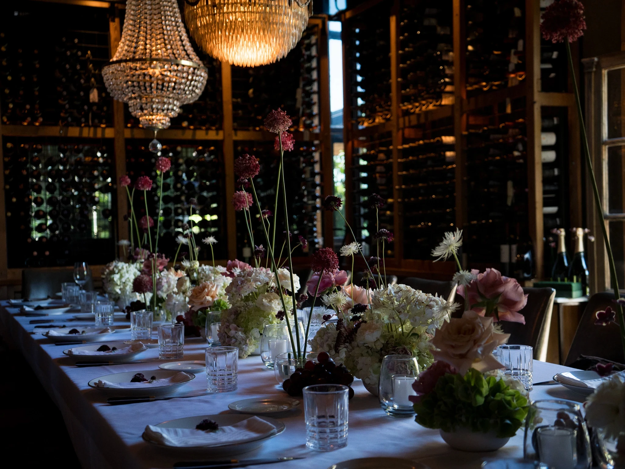 Salón de eventos con lámparas de cristal, mesas largas decoradas y paredes revestidas con botellas de vino.