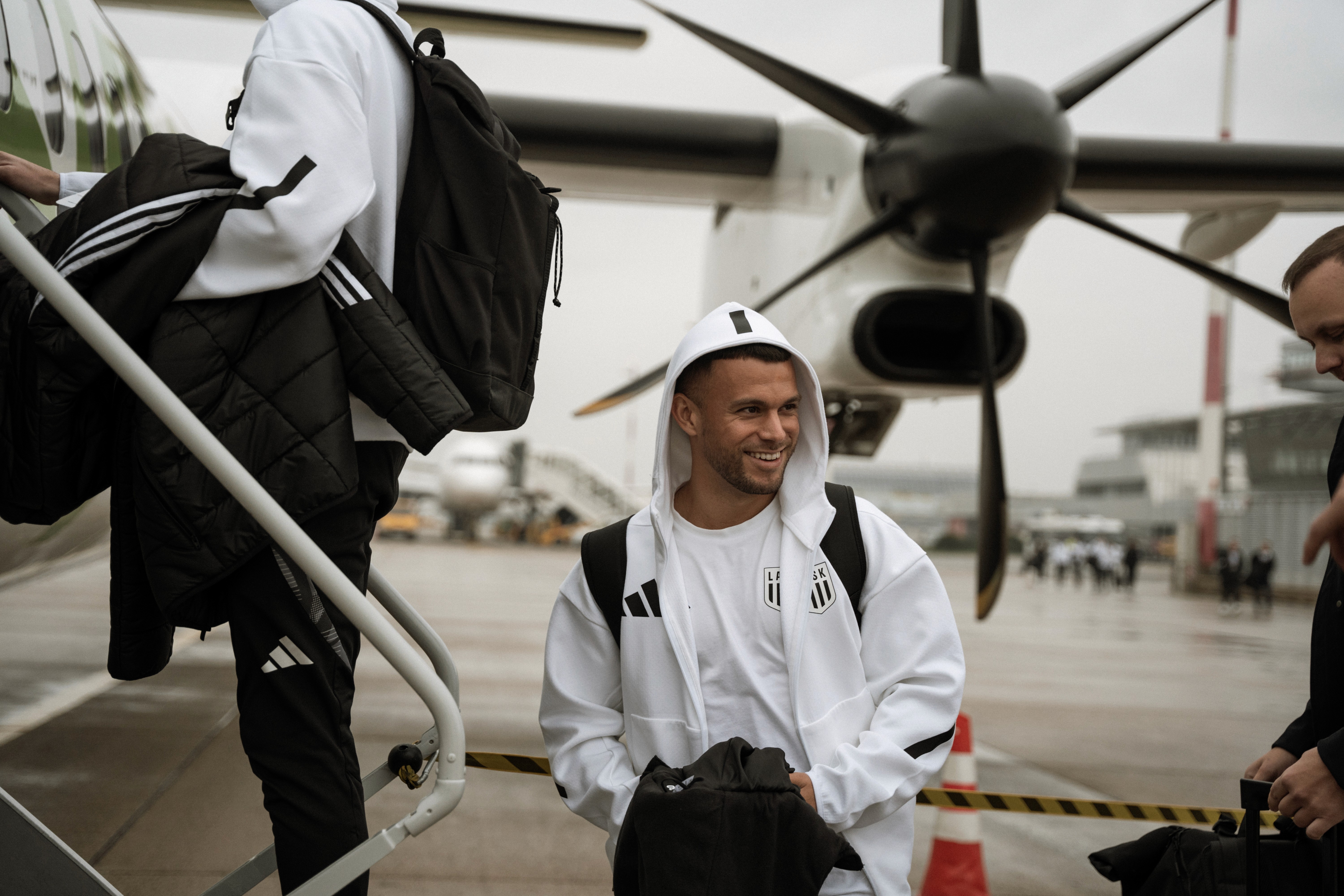 Ein lächelnder Mann in weißer Jacke und Kapuze steigt von einem Flugzeug. Er trägt eine schwarze Tasche und ist glücklich.