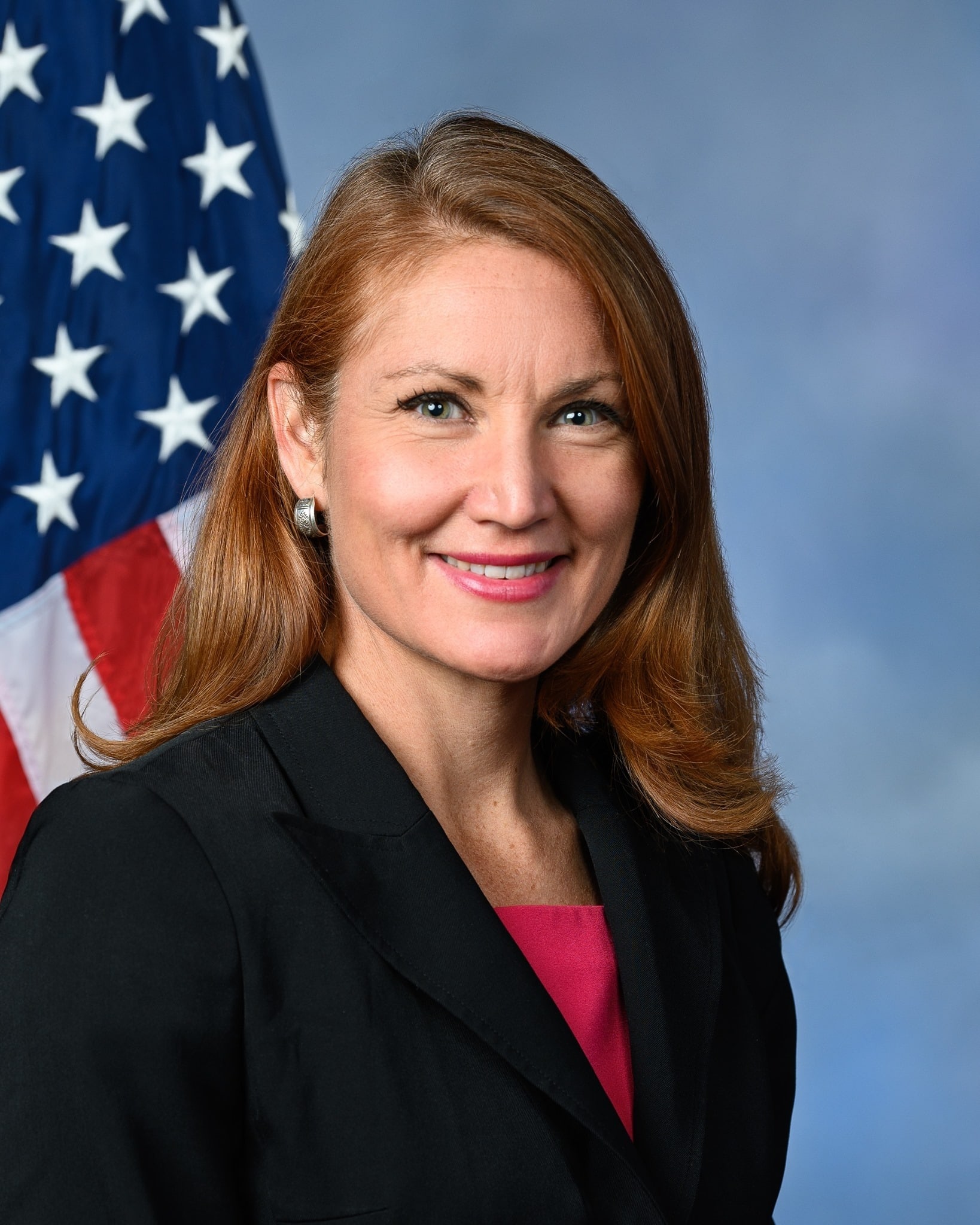 Portrait of Melanie A. Stansbury on a blue background with a portion of the flag of the United States of America in view behind subject