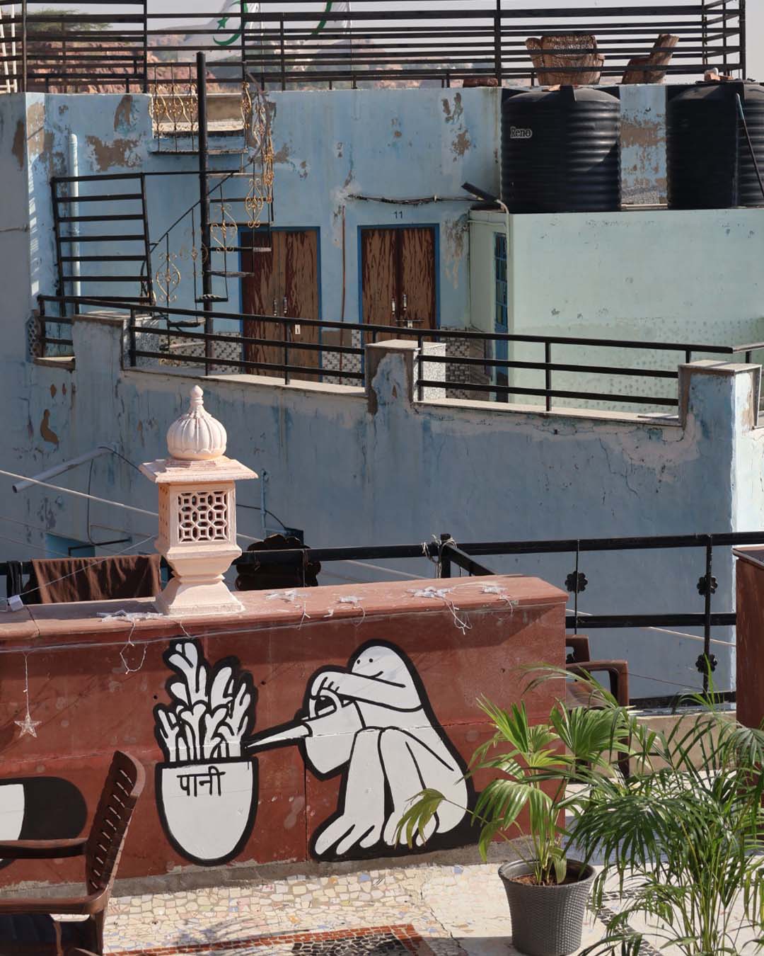 Mural painting of a joyful cartoon character watering a potted plant with the word ‘पानी’ written on it, painted black-and-white on a brown restaurant terrace with tables and oriental decorations visible.”