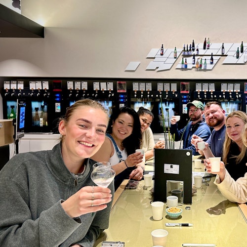 一組七個人微笑著，把杯子舉起，在一個光線明亮的房間裡圍著桌子，背景有架子和瓶子。