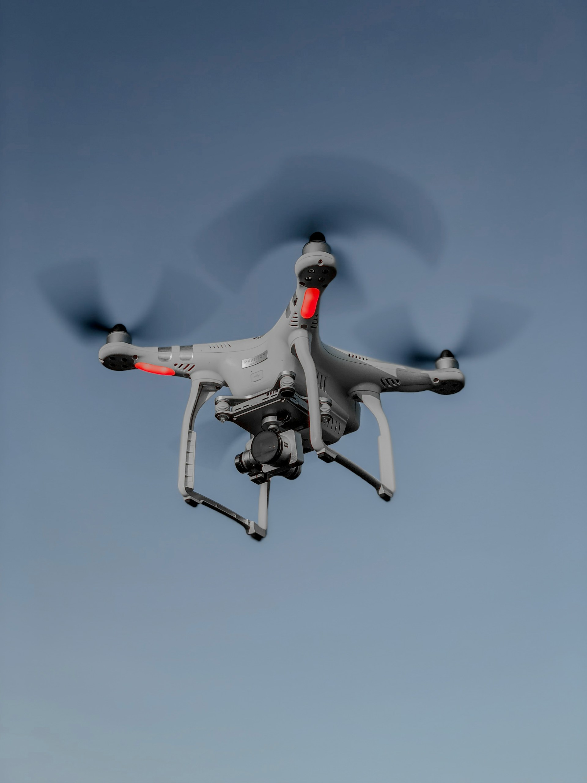 A white camera drone with spinning propellers flies against a blue sky.