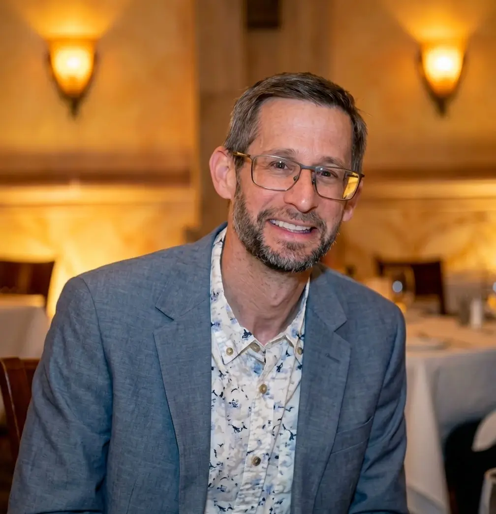 A man with a beard and glasses, wearing a light blue blazer over a patterned shirt, smiles warmly while seated at a restaurant with warmly lit ambiance in the background.