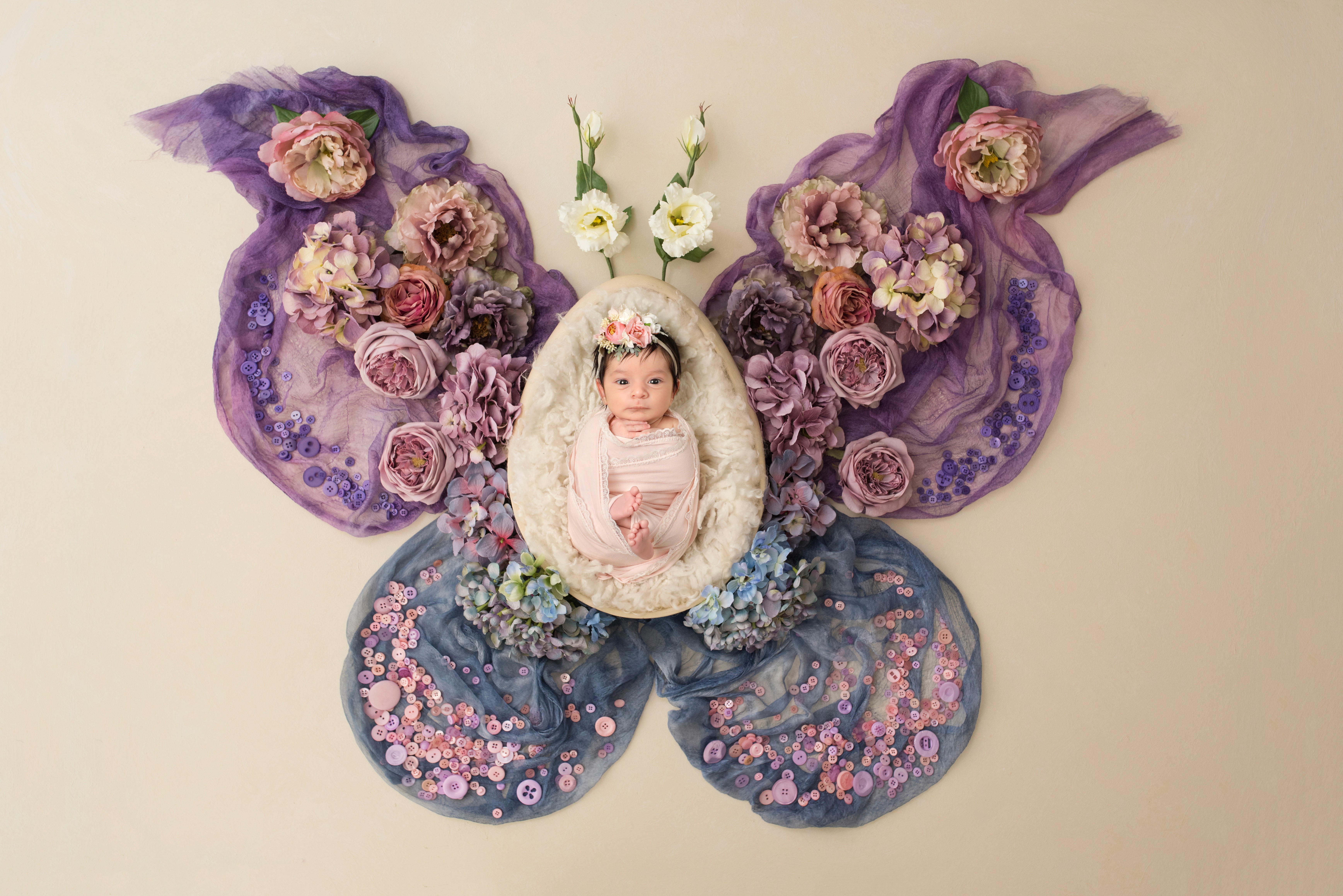 A newborn baby in a pink wrap lies in a nest centered within a large butterfly shape made of purple and blue flowers.