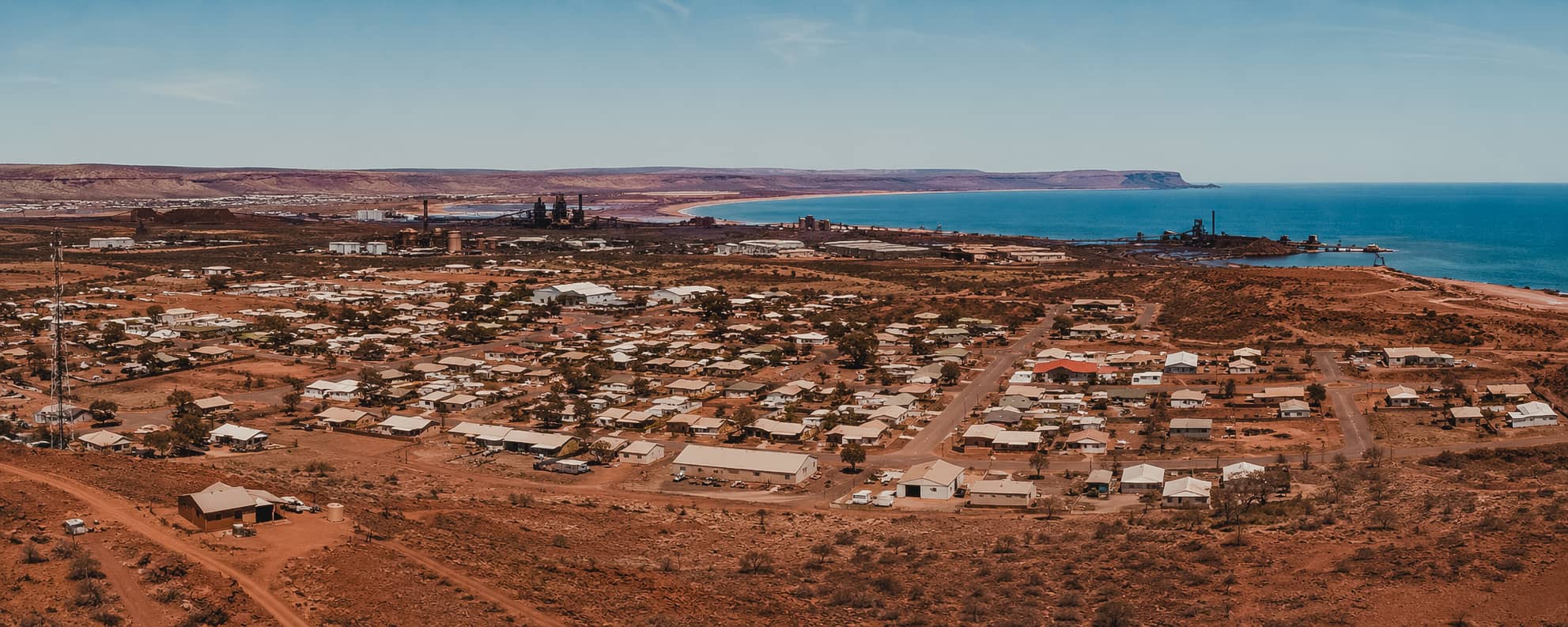 Aerial view of a remote Australian mining town with residential housing, industrial facilities, and the ocean in the background.