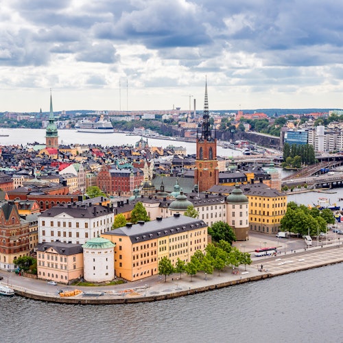 A panoramic view of a European city with colorful buildings, a prominent church tower, waterfront, and cloudy sky.