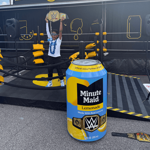 A child stands in a wrestling ring holding a championship belt overhead. In the foreground is a large, digitally added Minute Maid Lemonade can with WWE branding. The setup has yellow and black props and ropes.