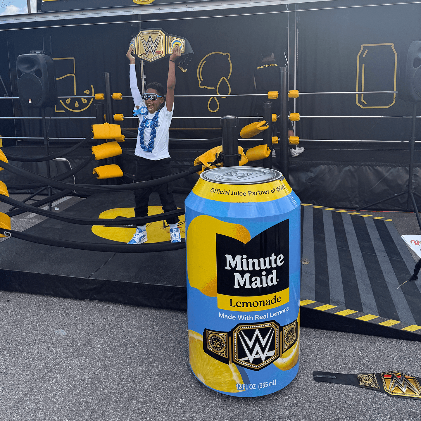 A child stands in a wrestling ring holding a championship belt overhead. In the foreground is a large, digitally added Minute Maid Lemonade can with WWE branding. The setup has yellow and black props and ropes.