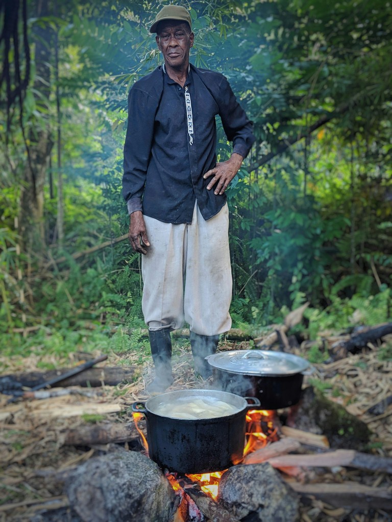 A man stands confidently in a lush forest setting, cooking over an open fire with large pots, capturing the essence of traditional outdoor cooking and self-reliance.