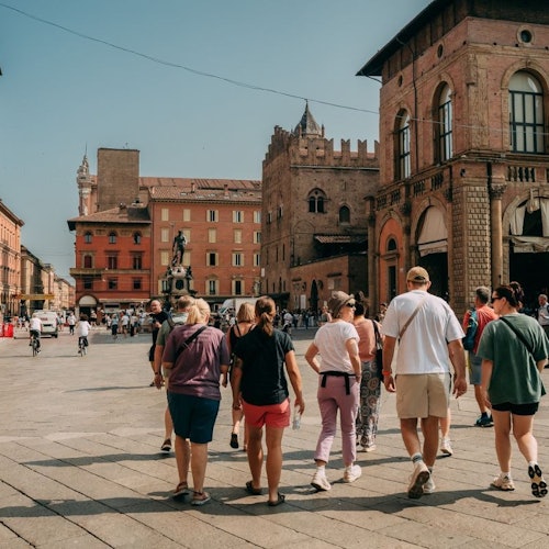 אנשים הולכים בכיכר עם בניינים היסטוריים, ופסל ברקע, ביום שטוף שמש.