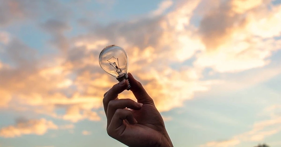 Hand holding a lightbulb in front of a sky with clouds.