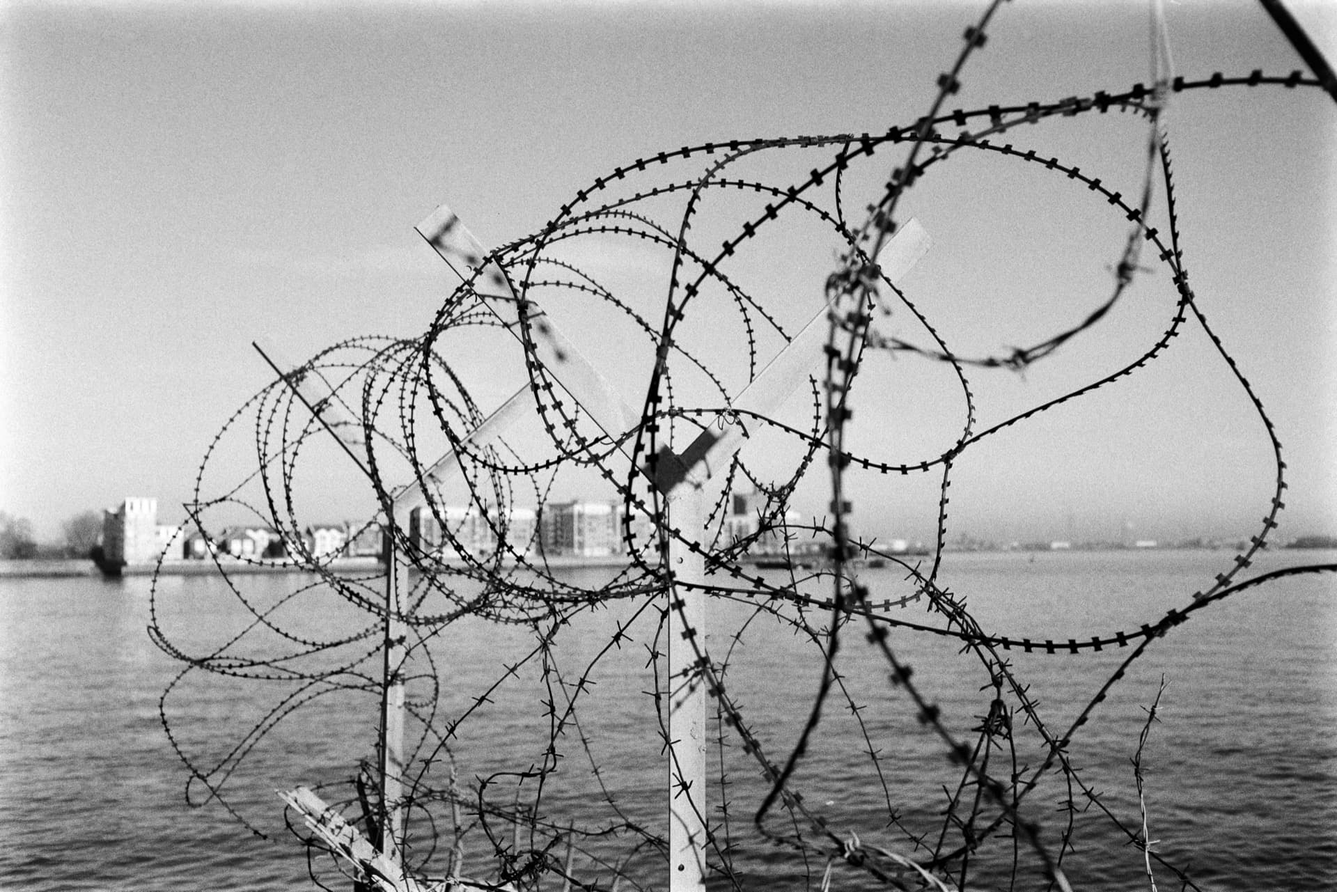 Coiled razor wire on chain-link fence across Thames, with industrial docklands and residential towers visible through winter haze beyond the water