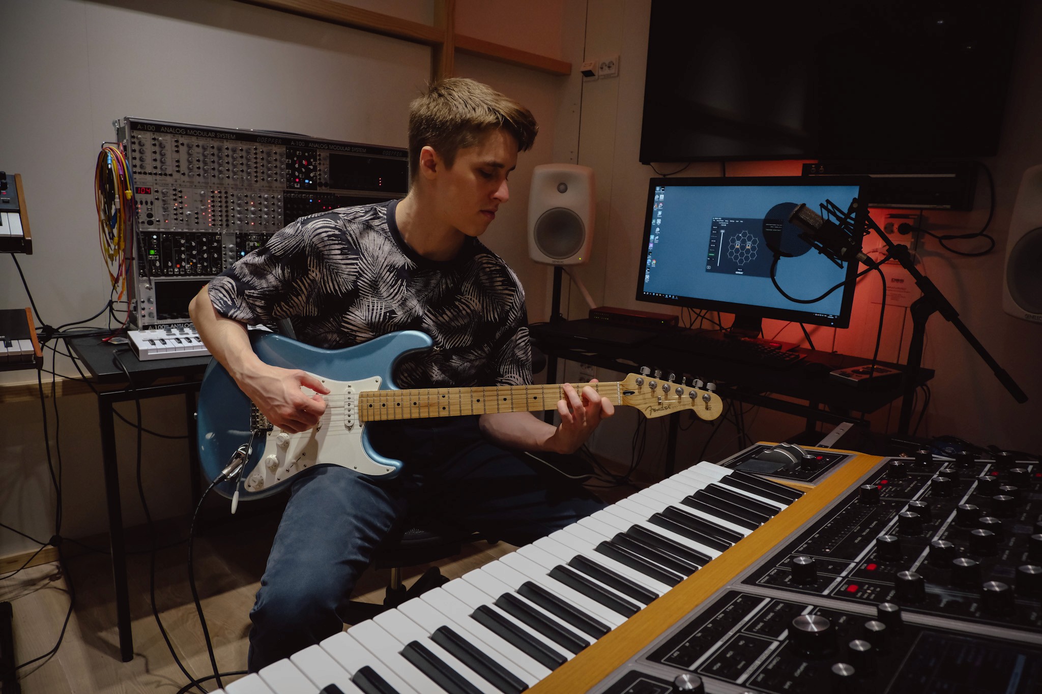 A musician sits at a keyboard, playing guitar, in a cozy recording studio filled with equipment.