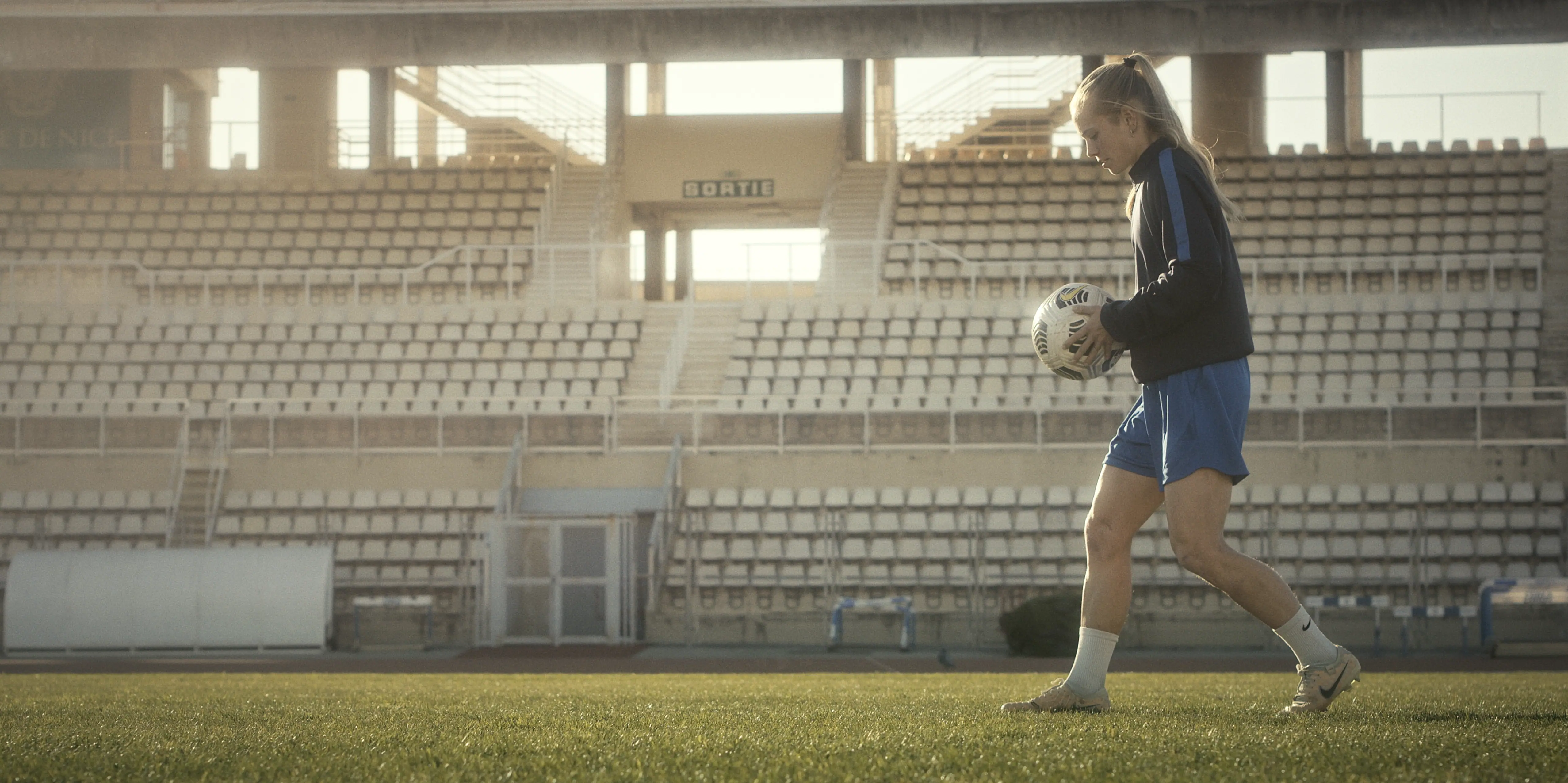 A woman in athletic attire dribbles a soccer ball on a sunlit field in an empty stadium, showcasing determination and focus during training.