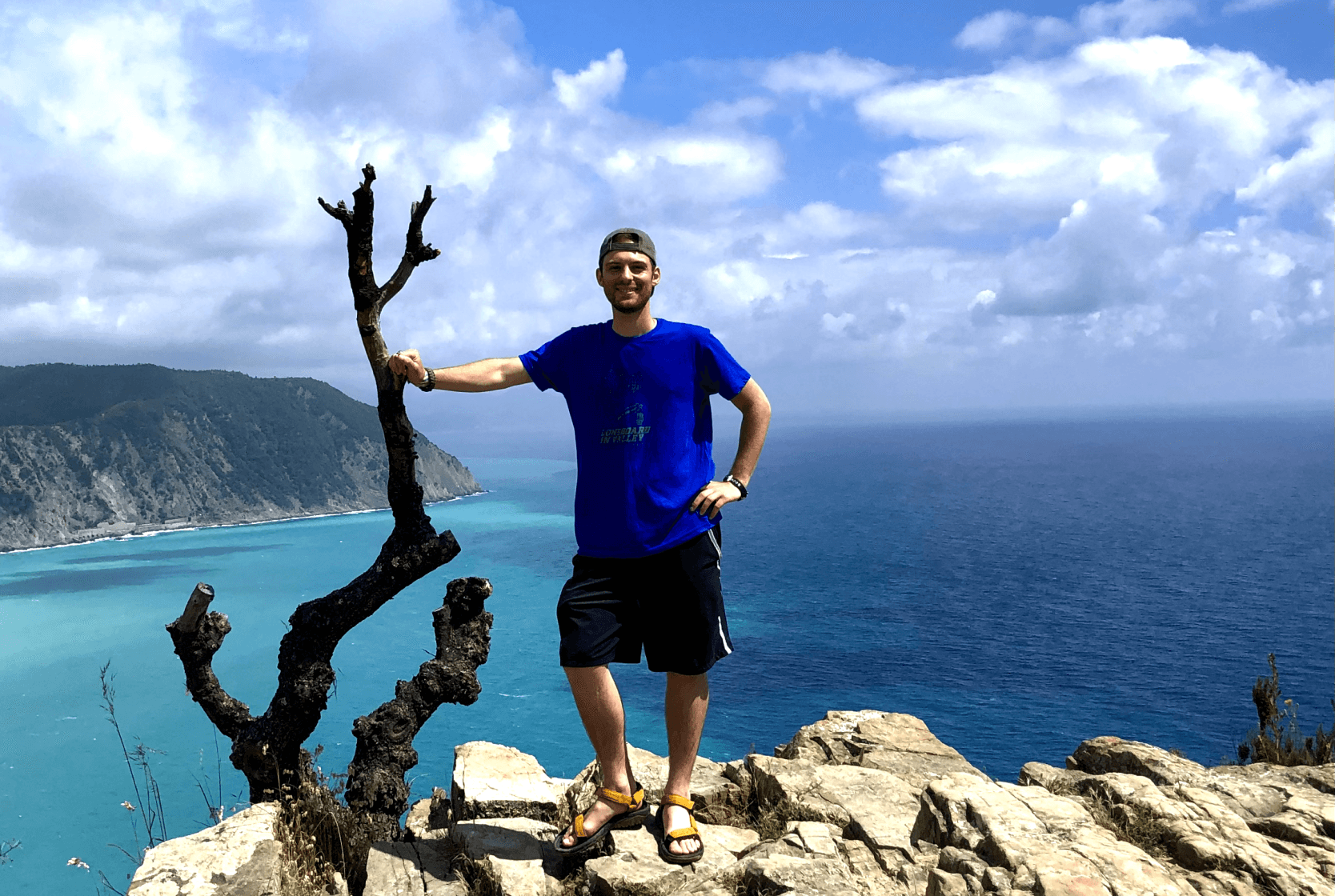 A man in a blue t-shirt and shorts stands on a rocky cliff with a scenic view of the ocean and distant cliffs in the background. He rests one arm on a bare, twisted tree, with a bright blue sky and clouds above.