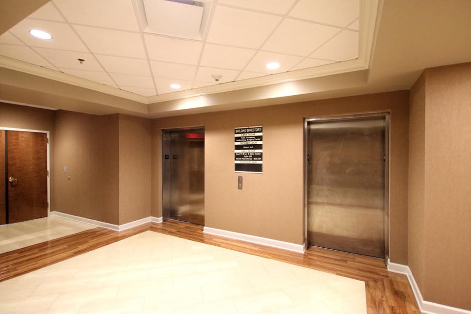 A spacious, well-lit room with beige walls, wooden flooring, and a small window with two elevator doors.