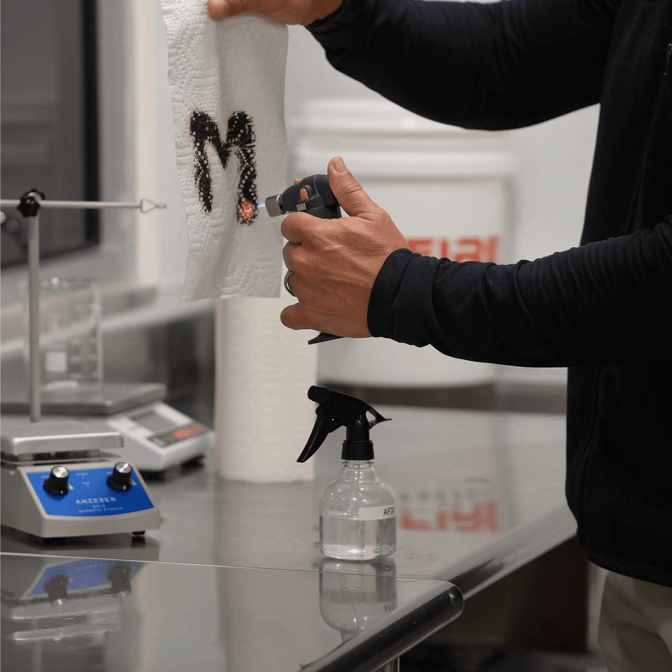 Close-up of a fire resistance test showing a blowtorch flame being applied in an M format to a treated paper towel in a laboratory setting.