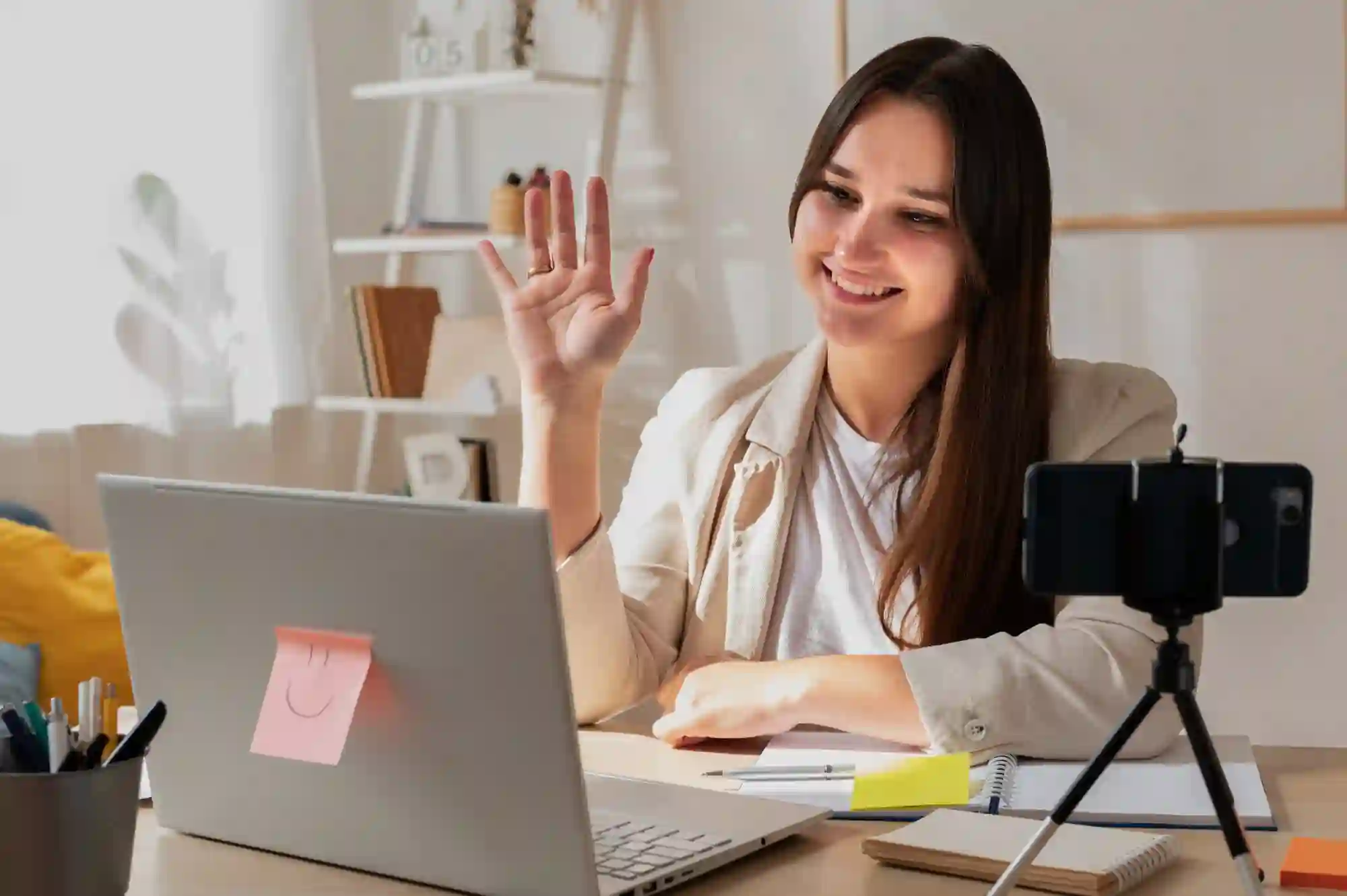 Woman smiling and waving during a video call on her laptop, with a smartphone on a tripod nearby.