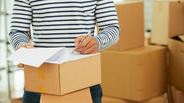 Homem de camiseta listrada azul e branca escrevendo em papéis sobre caixa de papelão em ambiente de estoque.