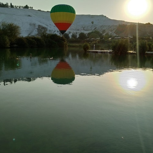 Isang hot air balloon ang lumulutang sa ibabaw ng tahimik na lawa na may bundok sa likuran sa pagsikat ng araw. Ang repleksyon ng lobo ay makikita sa tubig.