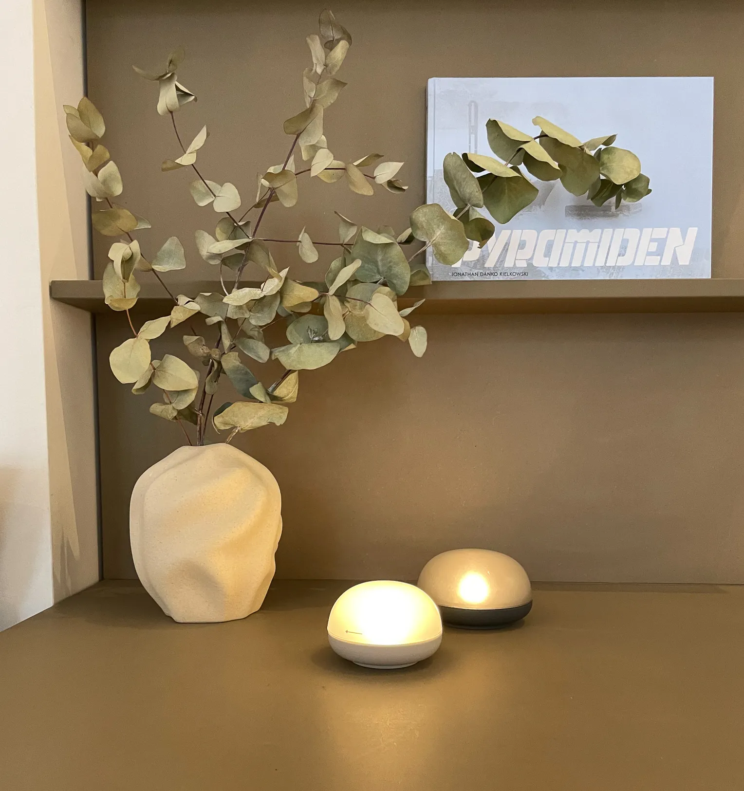Ein minimalistisches Regal präsentiert eine strukturierte beige Vase mit getrockneten Eukalyptuszweigen. Zwei kleine, leuchtende LED-Lichter spenden Wärme neben einem teilweise sichtbaren Buch.