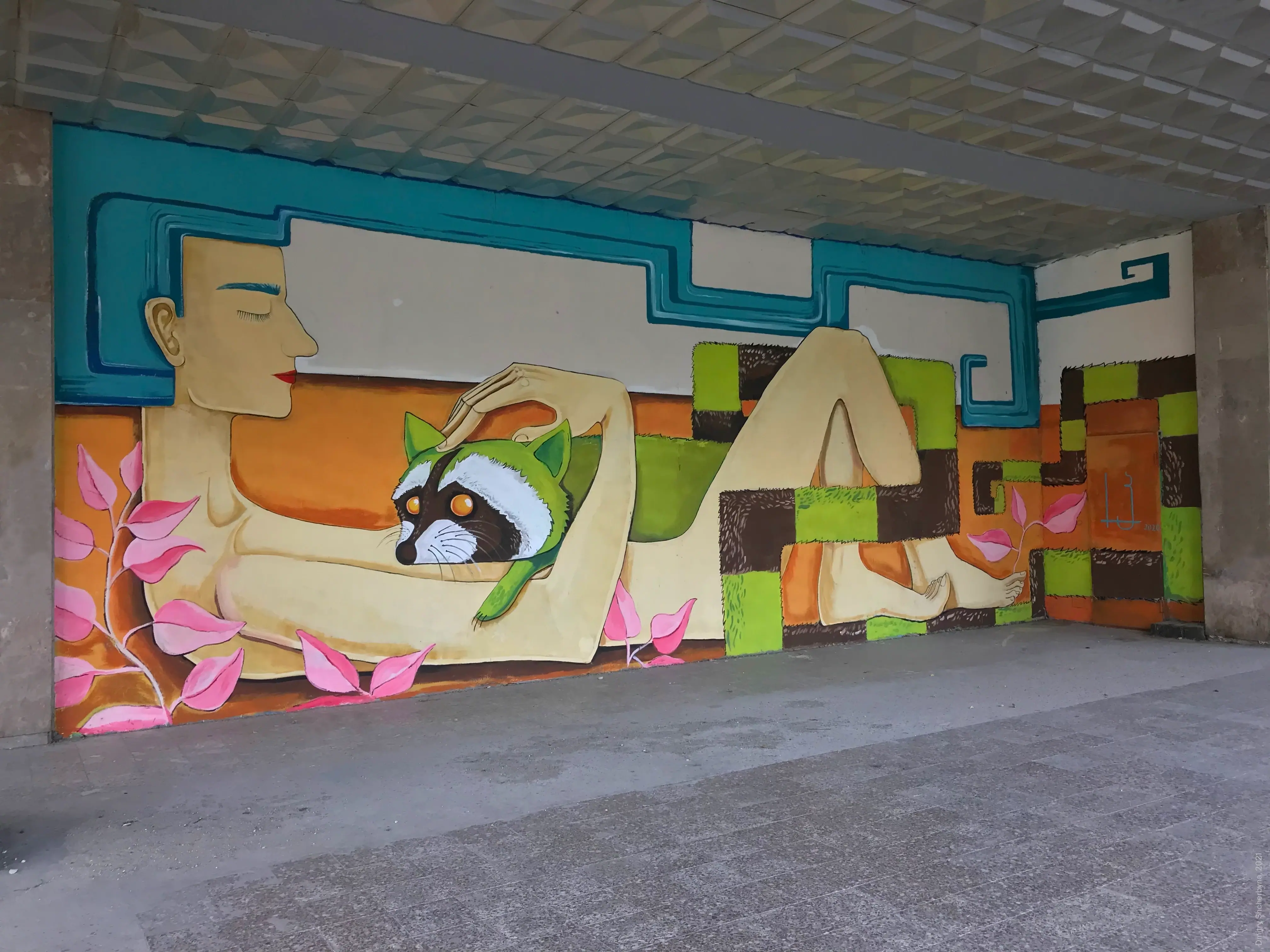 Mural of a stylized reclining figure with closed eyes, cradling a raccoon wearing a green hoodie, set against a background of colorful geometric blocks.