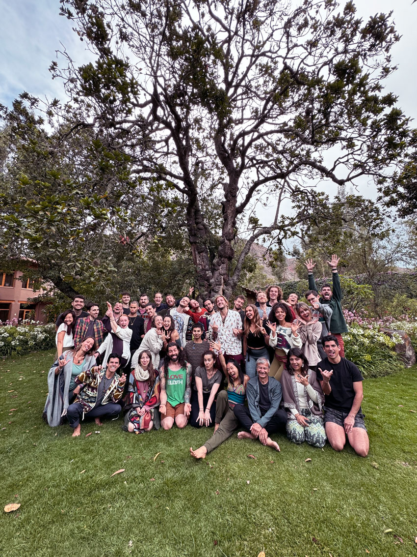 A large group of yoga teacher students smiling together on green grass, with a tall, leafy tree as a backdrop.