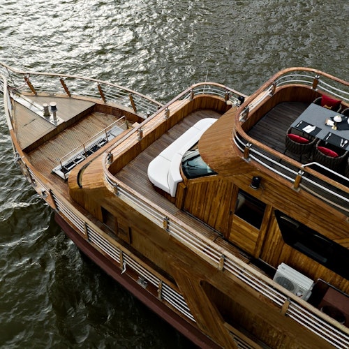 Un iaht de lemn pe apă, cu punți dotate cu balustrade, un loc de luat masa cu o masă și scaune roșii, și locuri de ședere în aer liber.