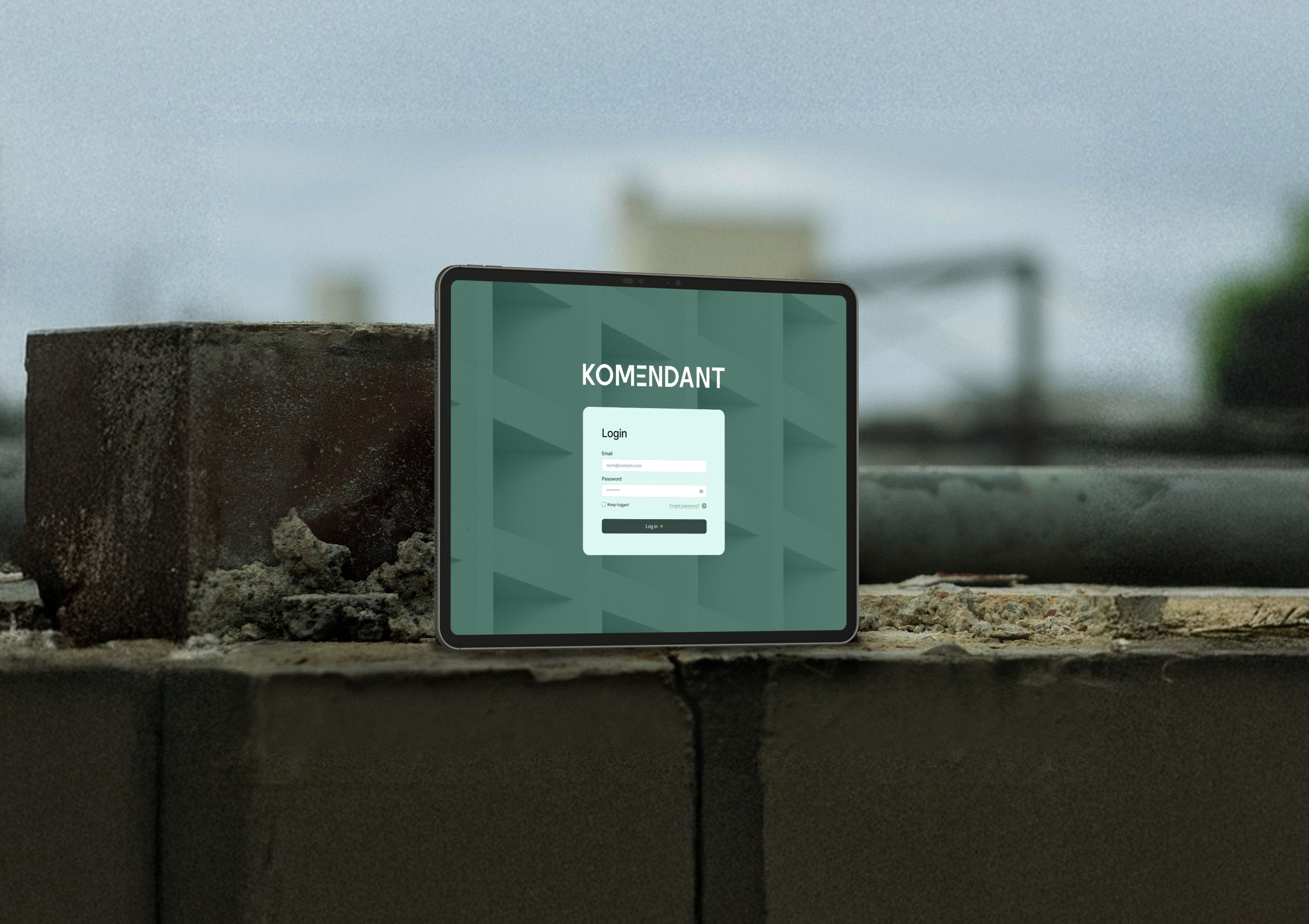 Tablet showing the KOMENDANT app login screen, placed at an angle on a perforated metal industrial surface.