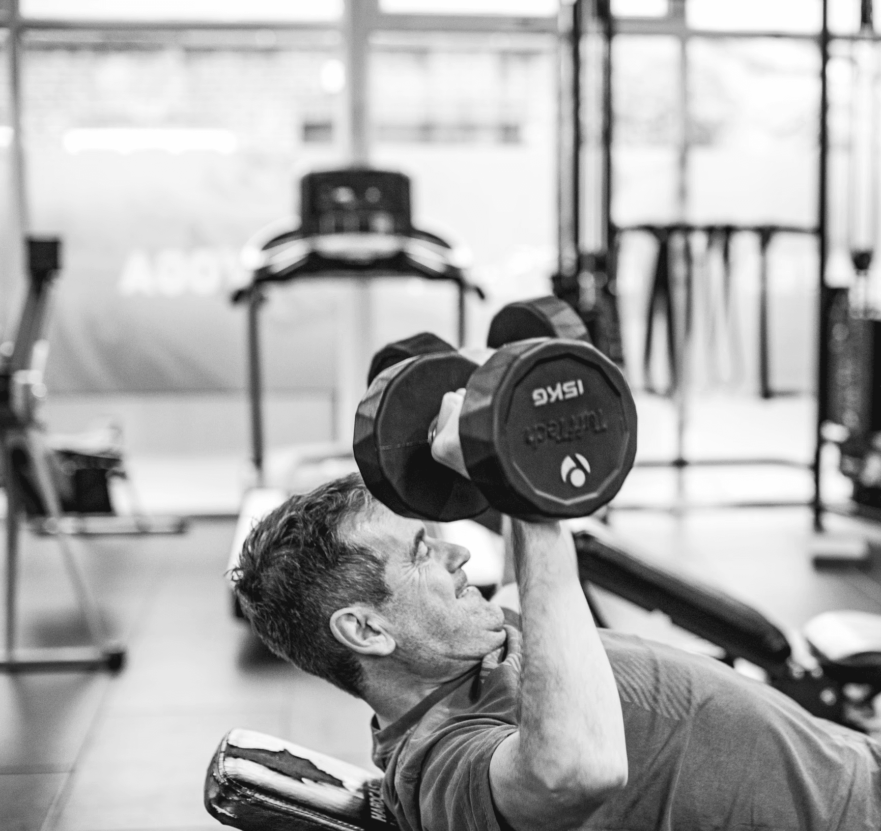 A man is performing a dumbbell chest press on a bench while another girl works out in the gym.