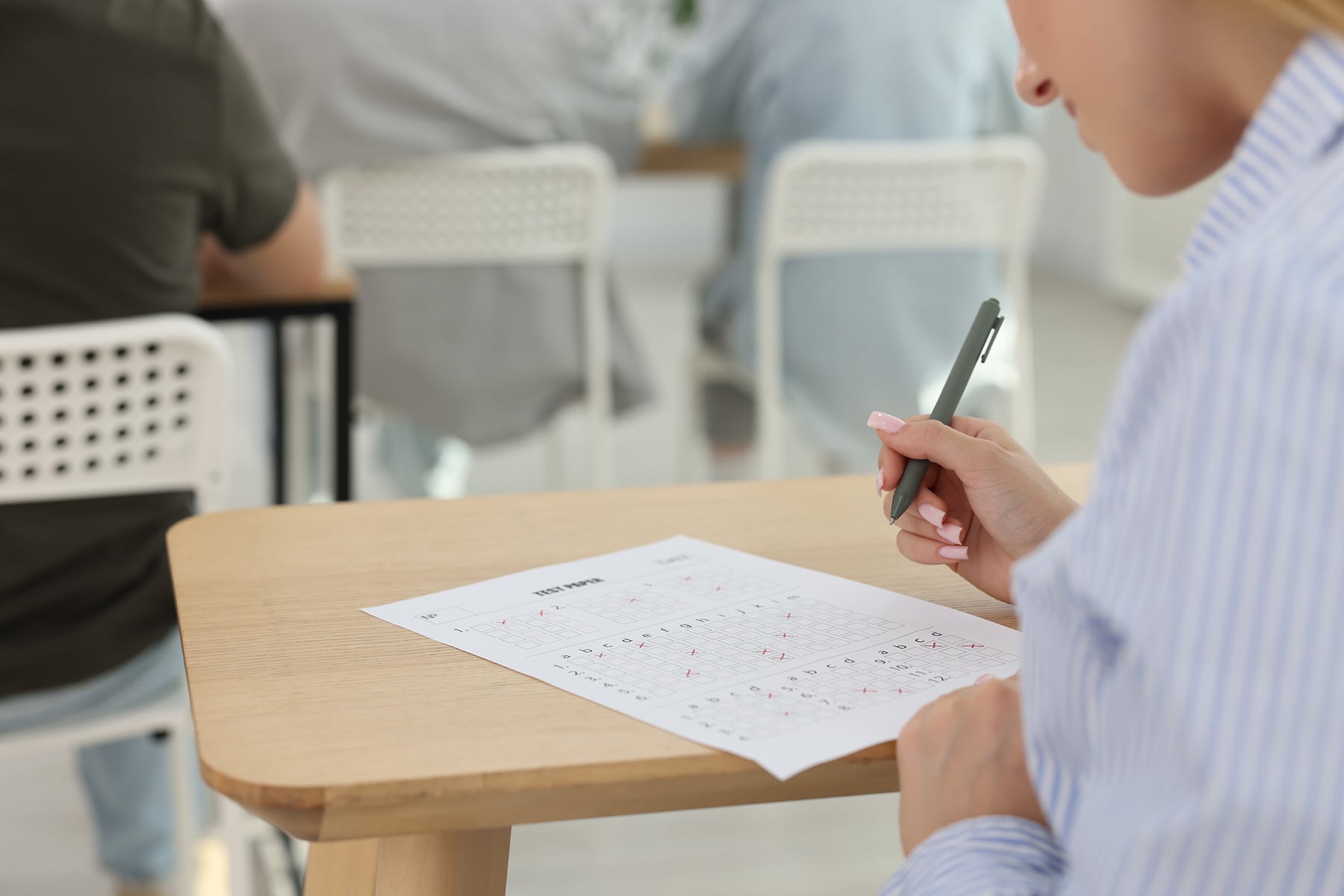 A student sits at a small desk completing a test paper, holding a pen while other students work in the background.