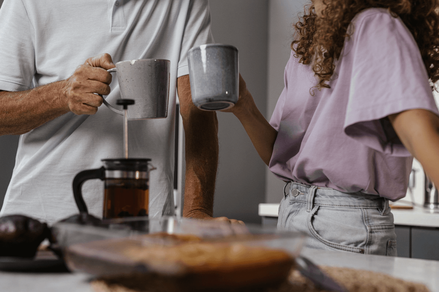 Two people standing at a kitchen counter in the morning, pouring coffee from a cafetiere into mugs.