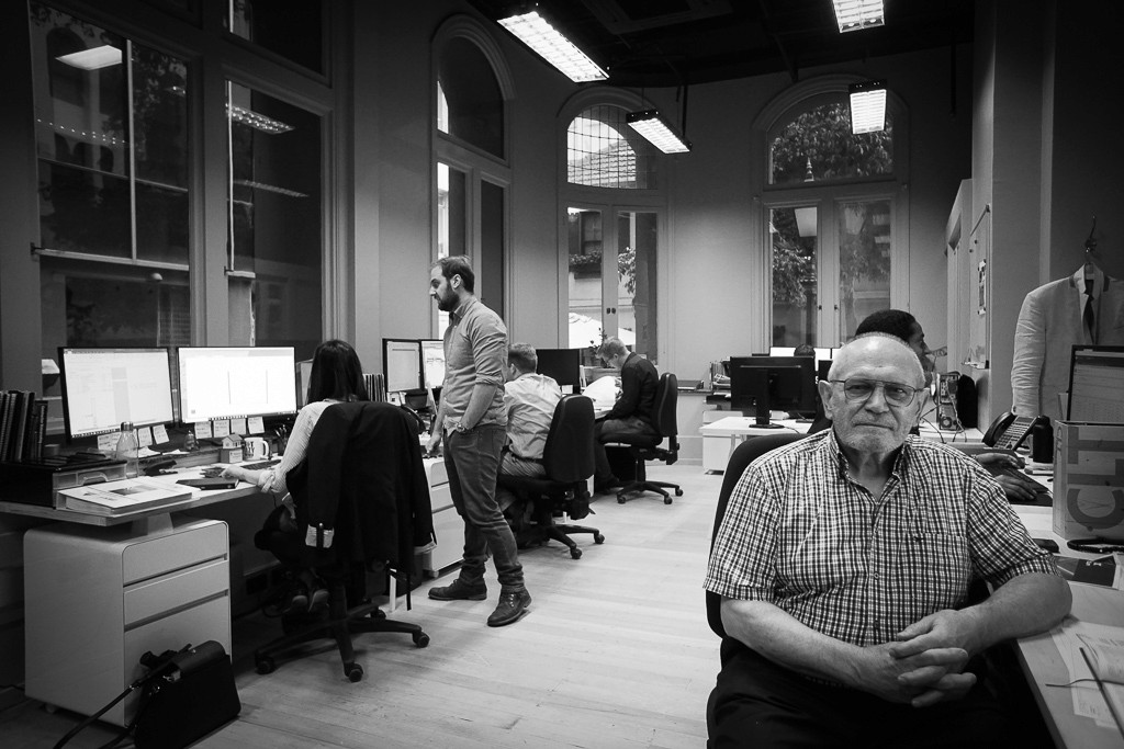 Black and white image of people working at a table with laptops, showing hands gesturing during discussion of digital content displayed on screens, with fabrica® logo in the corner.