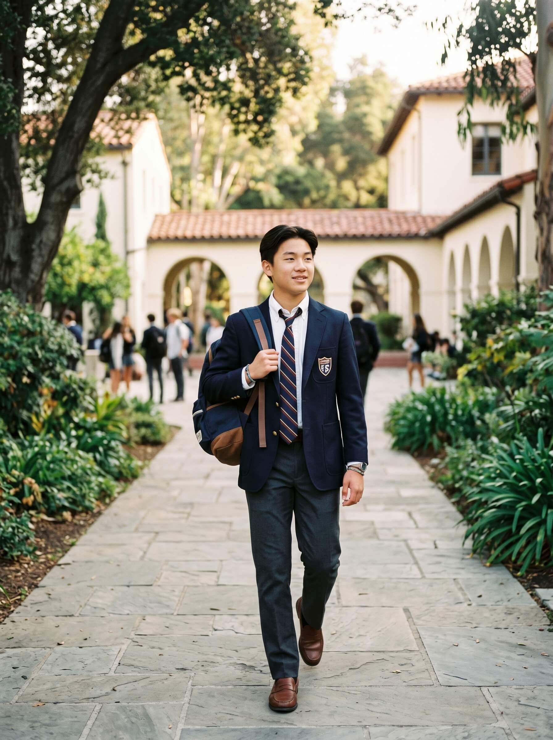 A young person in a navy school uniform with a tie and badge walks confidently along a stone path between two academic buildings, surrounded by green foliage and other students.